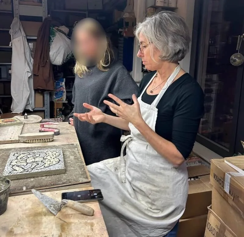 Two women in a workshop, one with gray hair wearing a white apron explaining something, and a younger woman with blurred face, listening. The workspace has tiles, tools, and boxes visible.