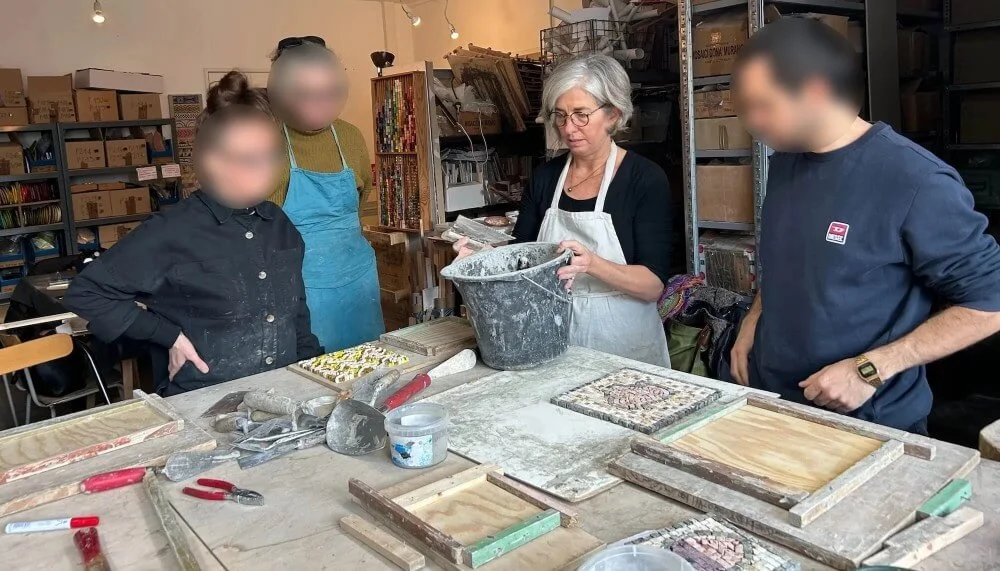 A group of four people gathered around a workshop table with various tools, brushes, and wooden frames, as an older woman demonstrates a craft process involving plaster or cement in a large container.