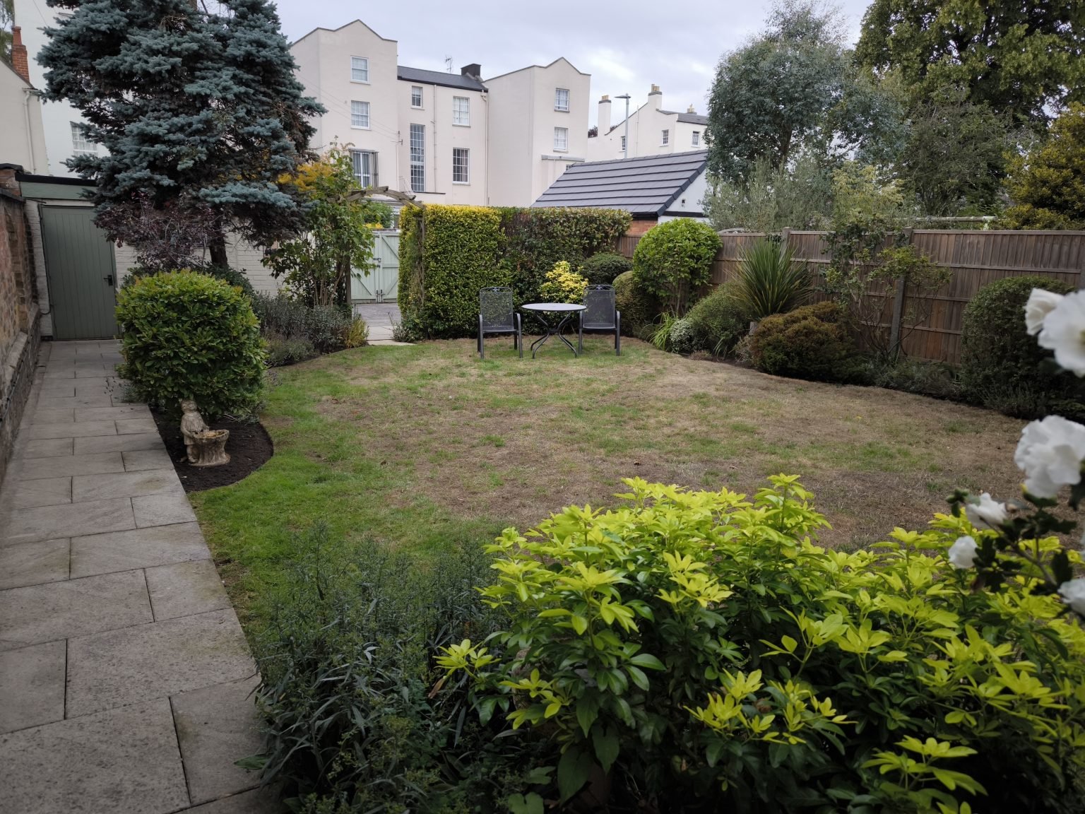 A backyard garden with a stone patio on the left, a lawn area in the center, and various shrubs, trees, and a small table with two chairs at the far end, enclosed by a wooden fence.