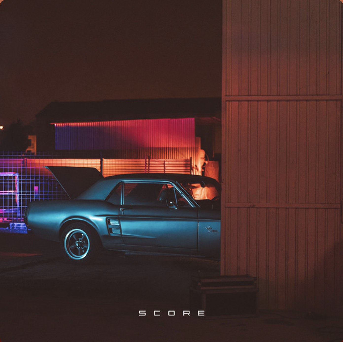 A vintage car parked near a red and orange warehouse, illuminated by neon lights in a dark, foggy setting, with the word 'SCORE' at the bottom.