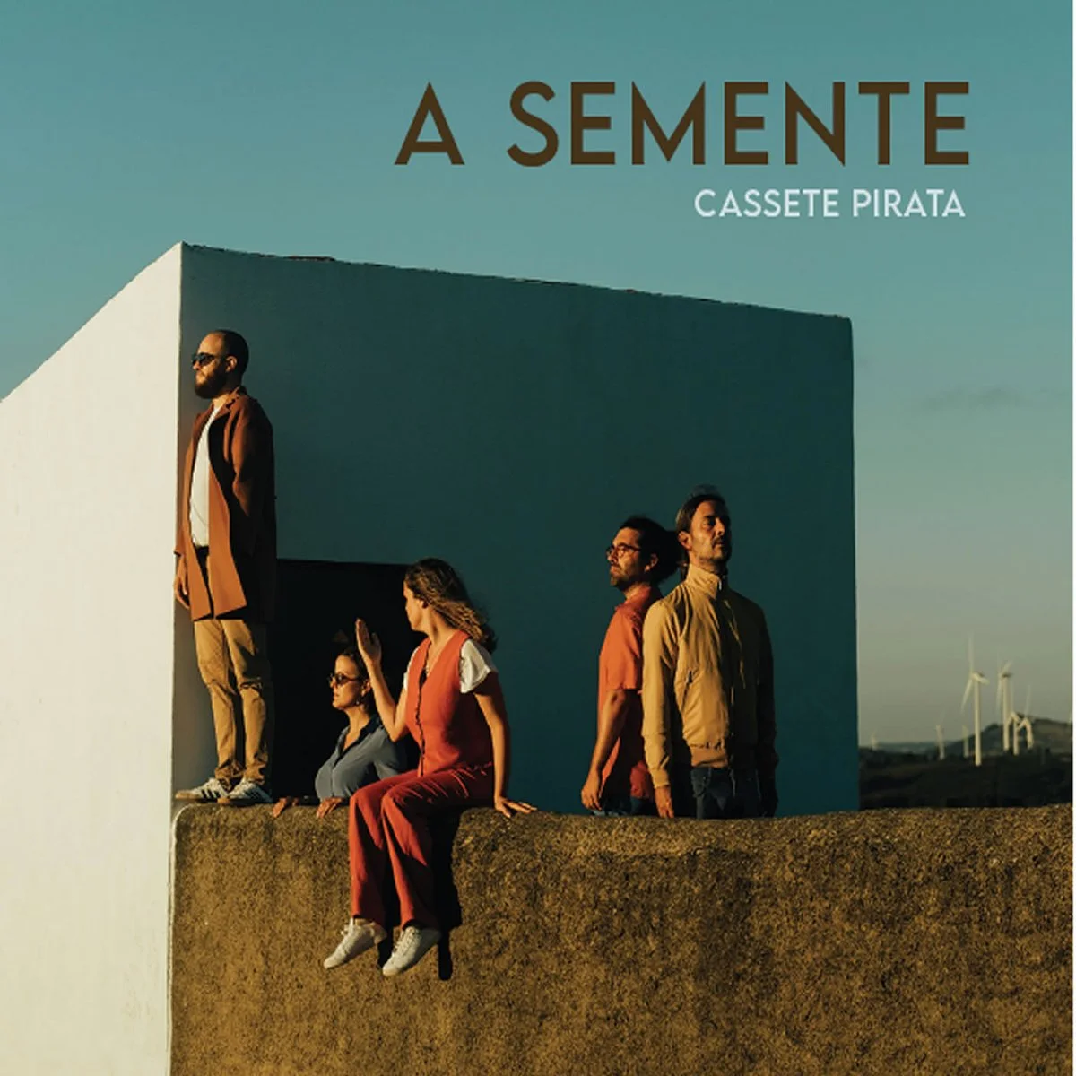 A group of five people, three men and two women, sitting and standing on a ledge outside a white modern building, with wind turbines in the background, during sunset.