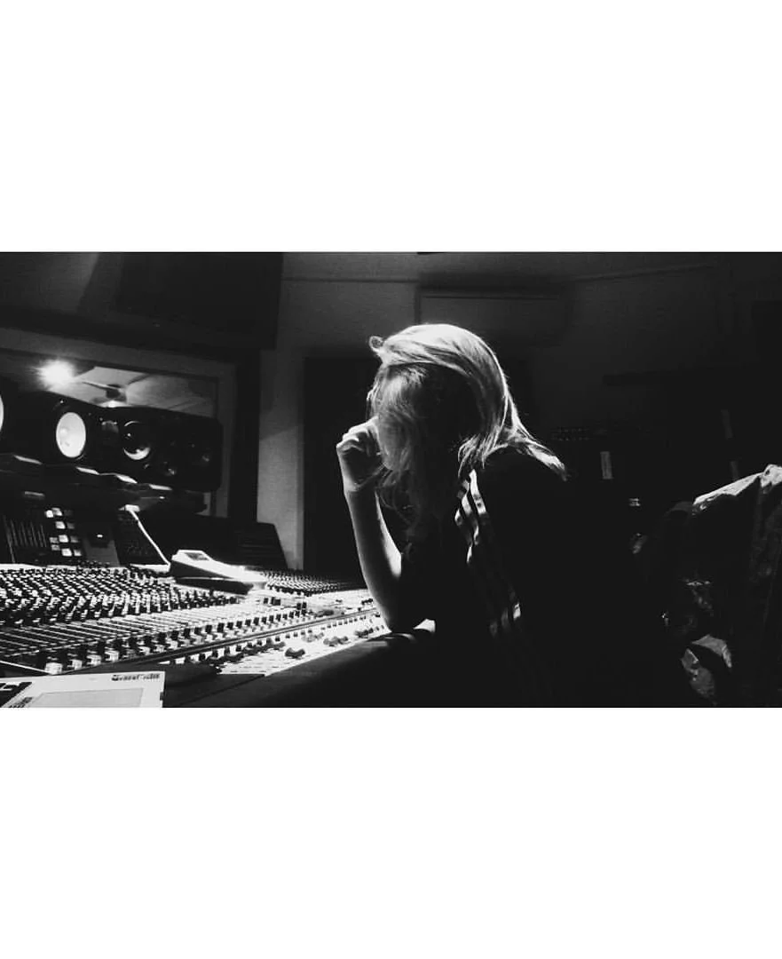 A woman with shoulder-length hair sitting at a mixing console in a recording studio, resting her head on her hand, with studio speakers visible in the background.