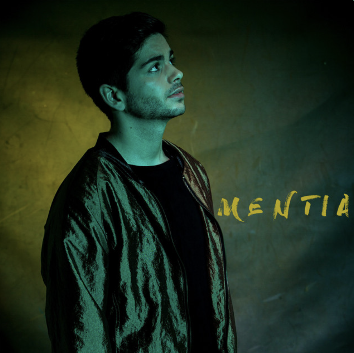 A young man with dark hair and stubble looking upwards, wearing a shiny black jacket, against a yellow-green background with partially visible yellow text.