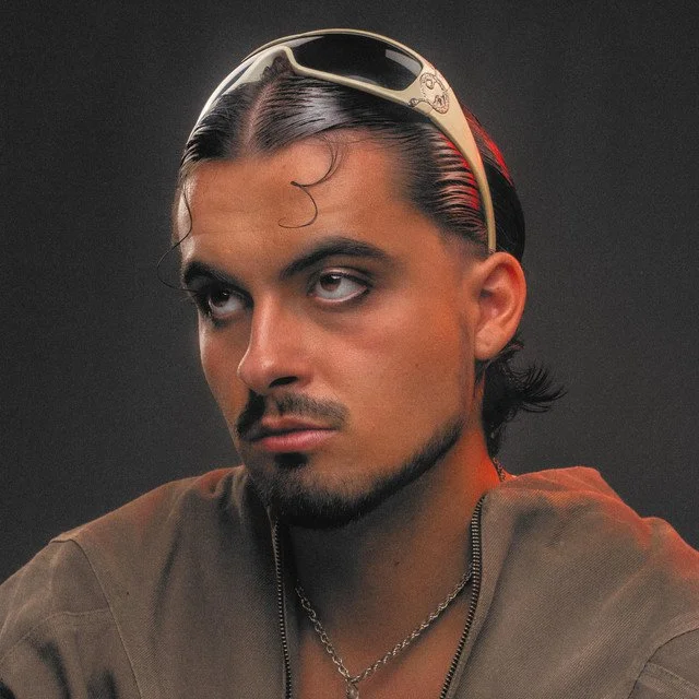Close-up of a man with dark hair, fierce expression, wearing sunglasses on head, a brown shirt, and a silver chain necklace.