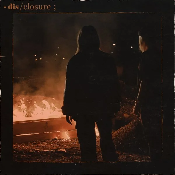 Silhouette of a person standing in front of a large fire at night, with another person nearby, both surrounded by dark environment.