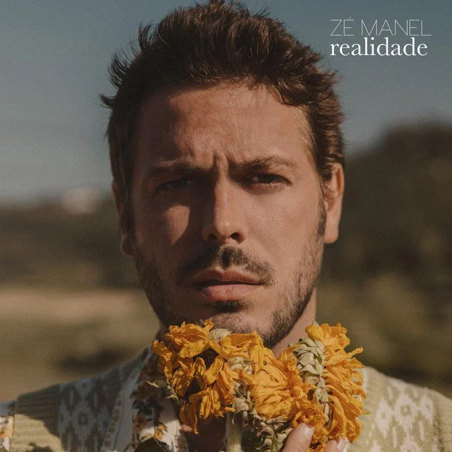 Close-up of a man with a serious expression, wearing a sweater with a flower garland around his neck, outdoors with a blurred background.