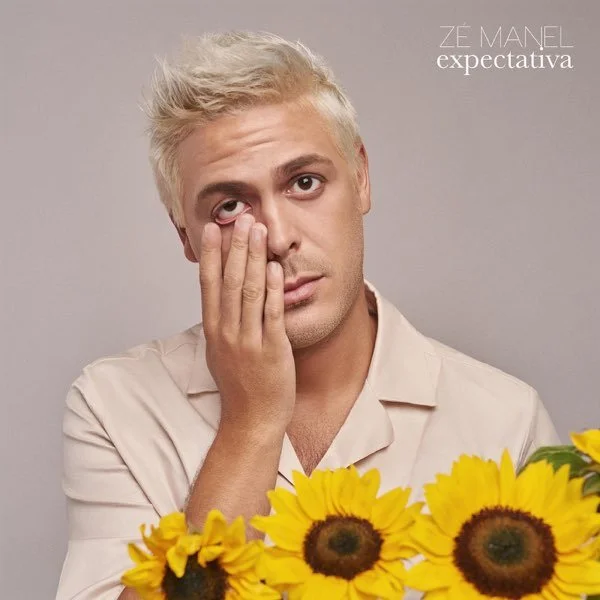 A man with platinum blonde hair resting his face on his hand, looking tired or contemplative, with a bouquet of sunflowers in front of him and a plain gray background. Text in the top right corner reads "ZÉ MANEL" and "expectativa."