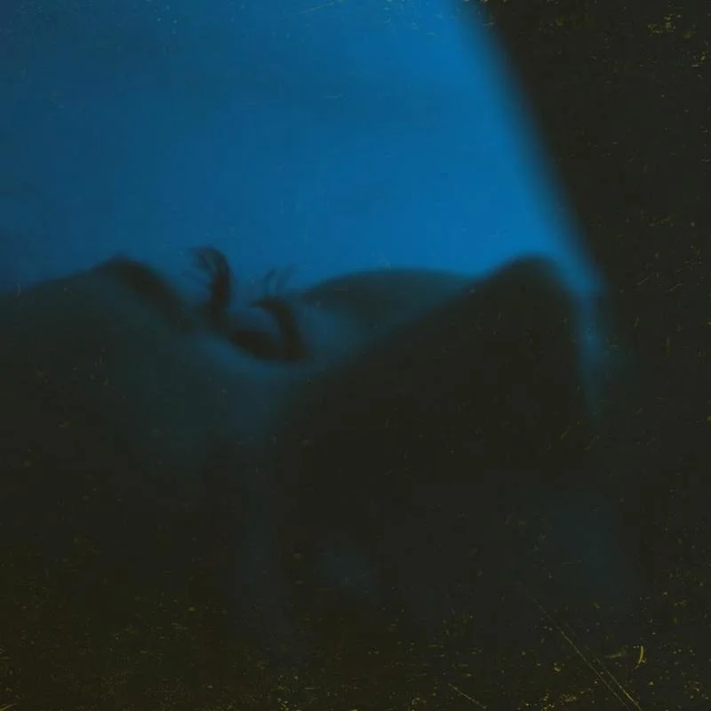 Close-up of a sleeping person's face with eyes closed and head resting on a surface, illuminated by soft blue light.