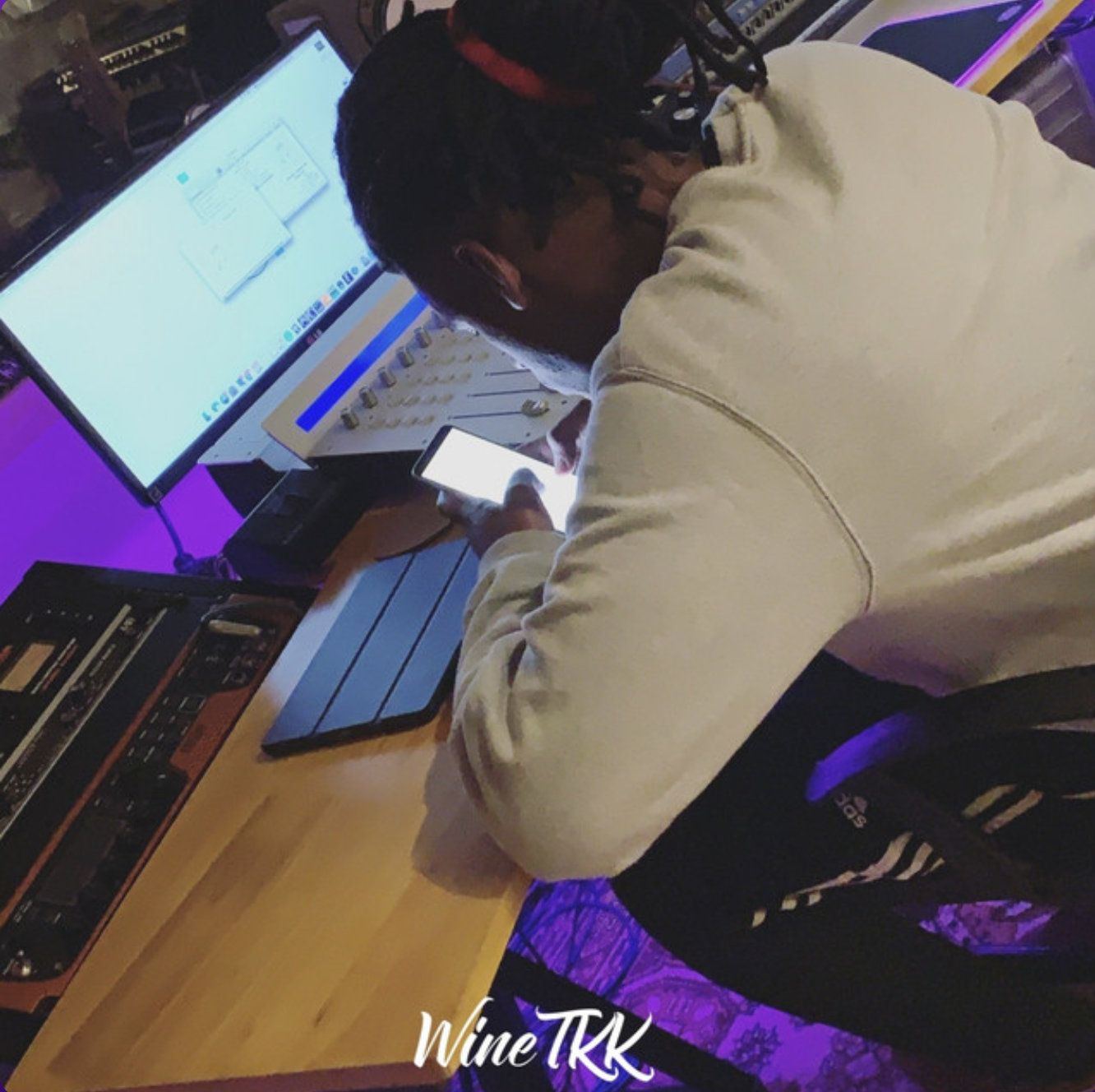 A person with braided hair, dressed in a white hoodie, sitting at a wooden table with electronic music equipment, working on a computer and looking at a smartphone.