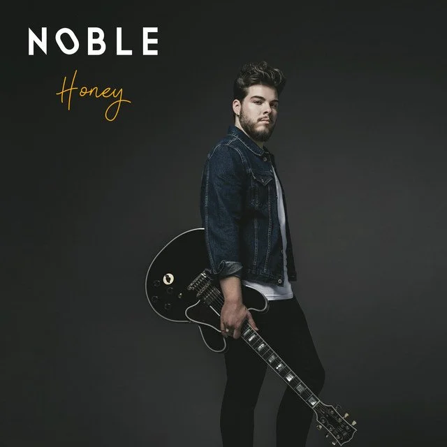 A young man with a beard and styled hair holding a black electric guitar, standing against a plain dark background. The text 'Noble Honey' is displayed in the top left corner.