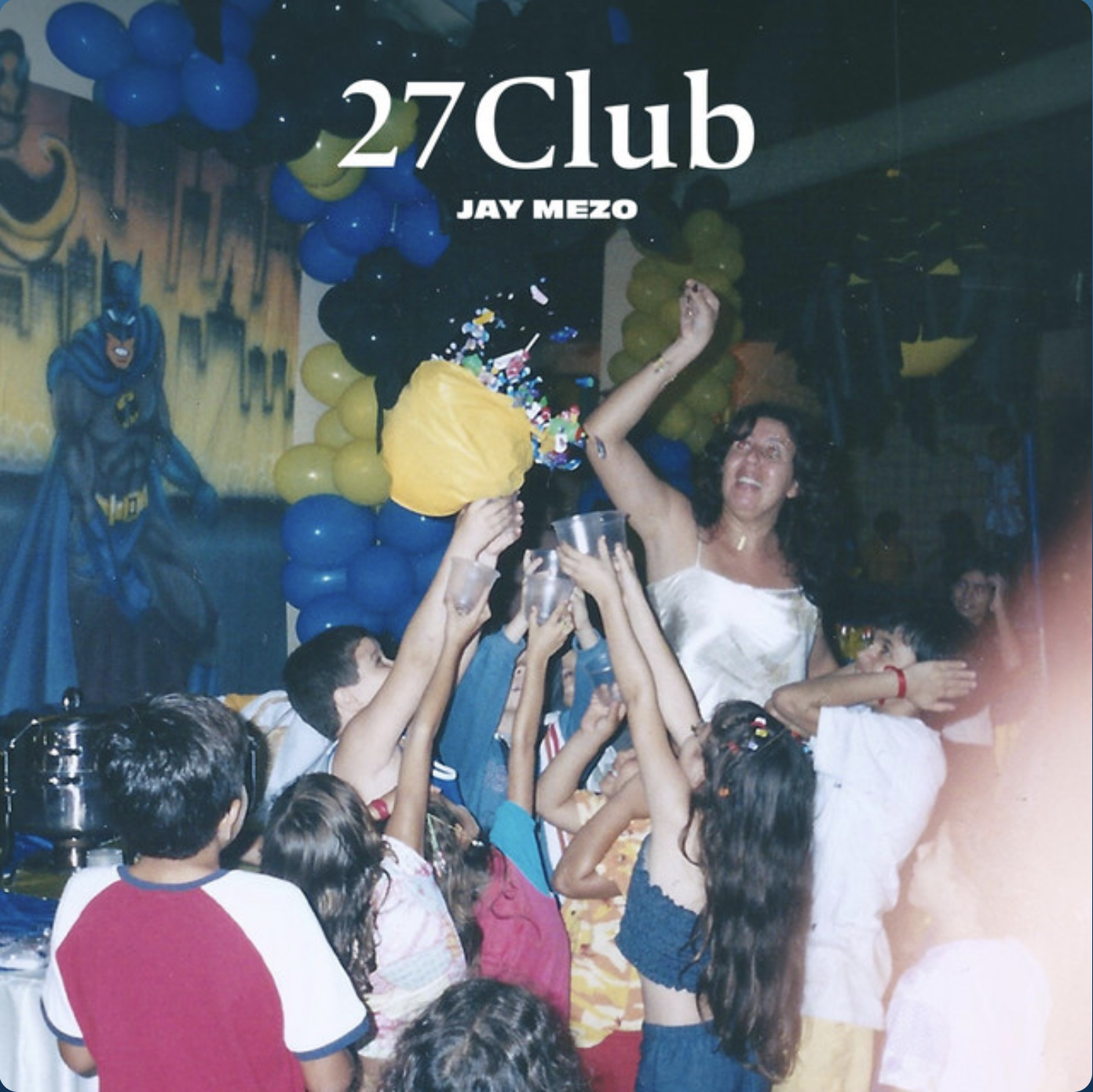 Children celebrating a birthday party with a woman, possibly the birthday girl, holding a balloon bouquet, surrounded by kids reaching for gifts or prize, with a Batman poster and festive yellow and blue balloons in the background.