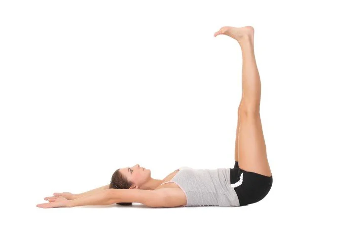 white woman in shorts and a tank top lying on her back, her arms overhead, and legs reaching upwards.