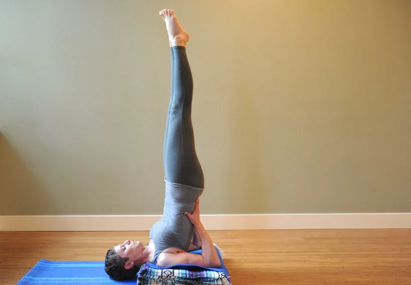 white woman with short hair practicing Salamba Sarvangasana (shoulderstand) on a stack of Mexican folded blankets