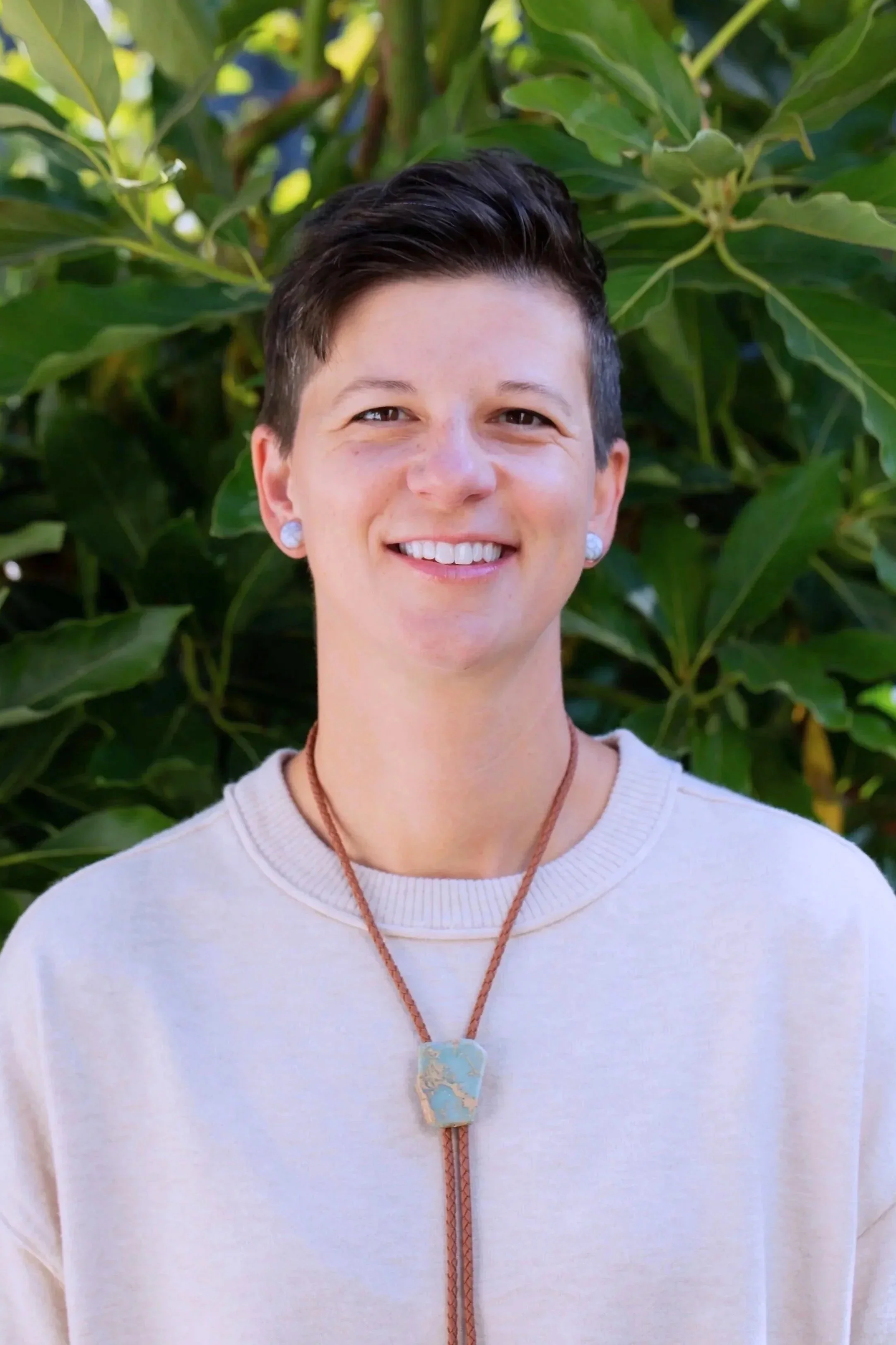 Close-up of a smiling woman with short dark hair and earrings, wearing a beige sweater and a necklace with a large, multicolored pendant, standing outdoors in front of green leafy plants.