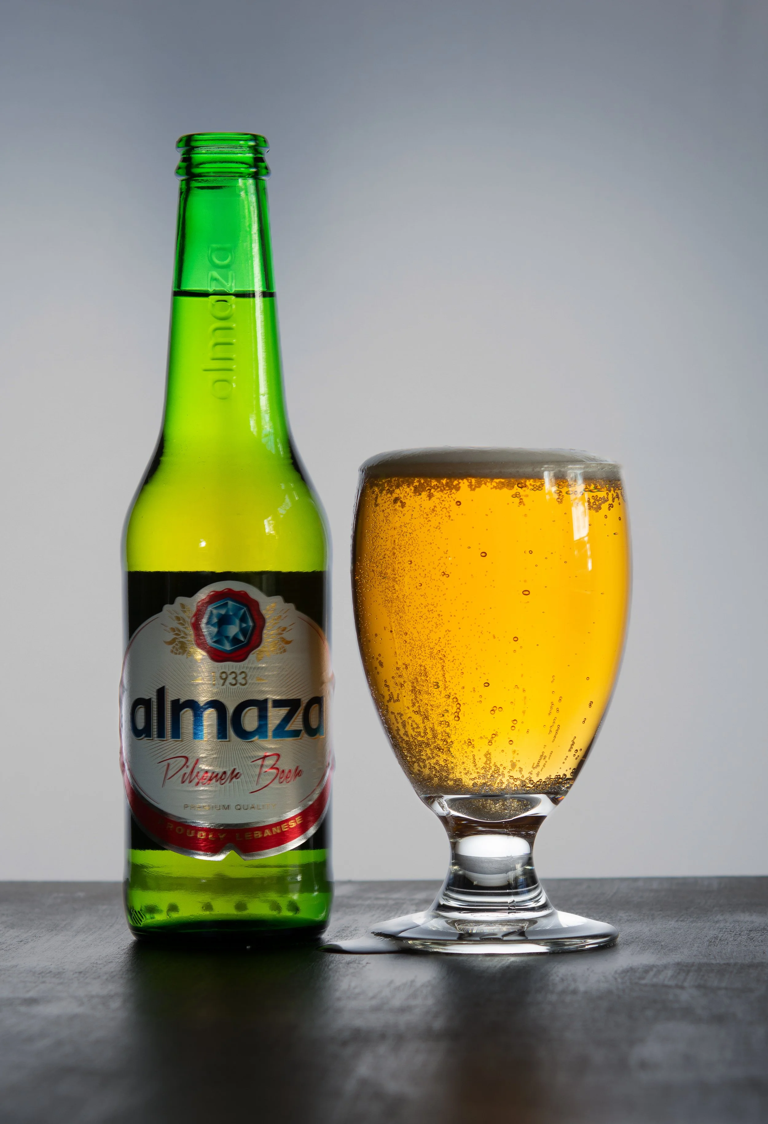 Une bouteille de bière Almaz et un verre rempli de bière blonde, placés sur une surface en bois avec un fond gris clair.
