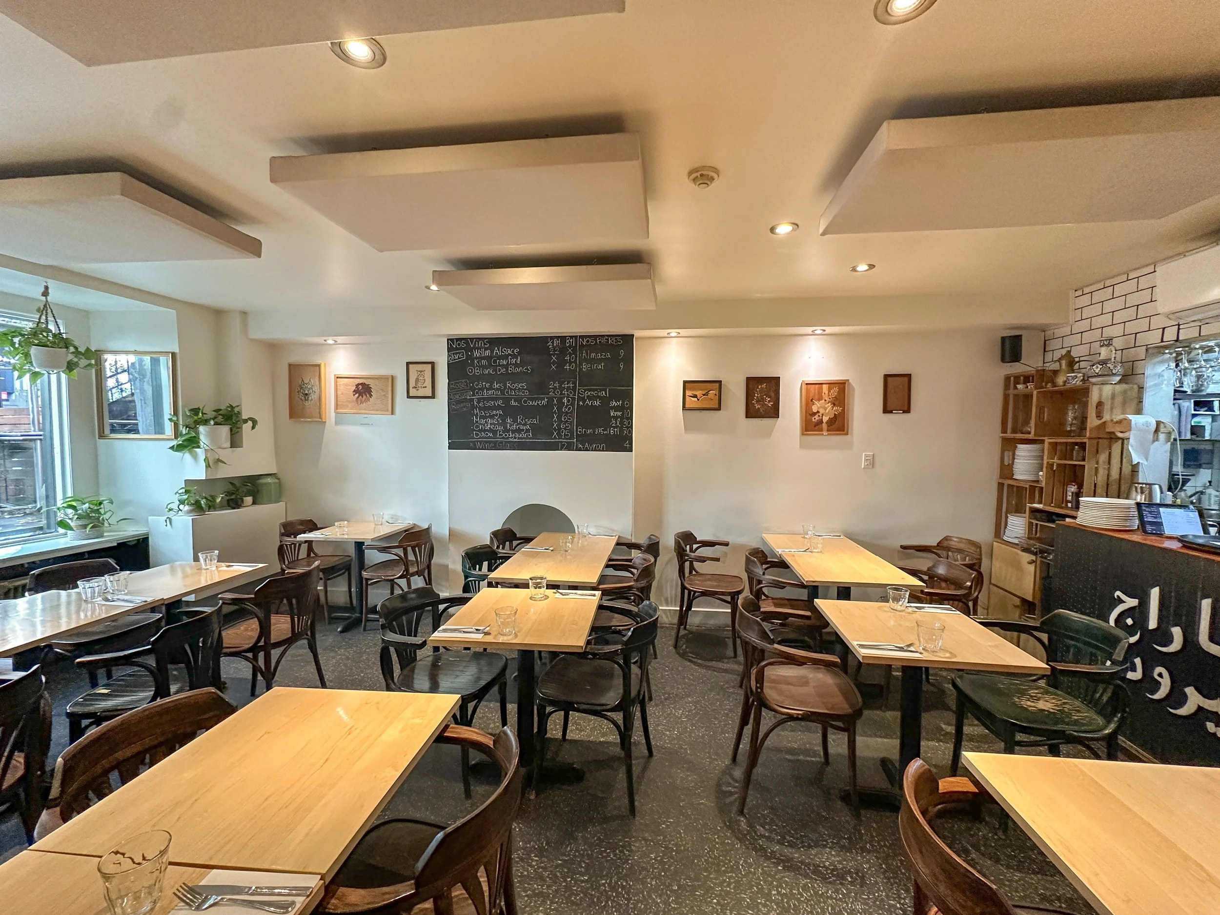 Salle à manger d'un restaurant avec plusieurs tables en bois, des chaises en bois et en plastique, des plantes vertes près des fenêtres, un tableau noir avec le menu, des œuvres d'art suspendues sur le mur, une cuisine partiellement visible à droite.