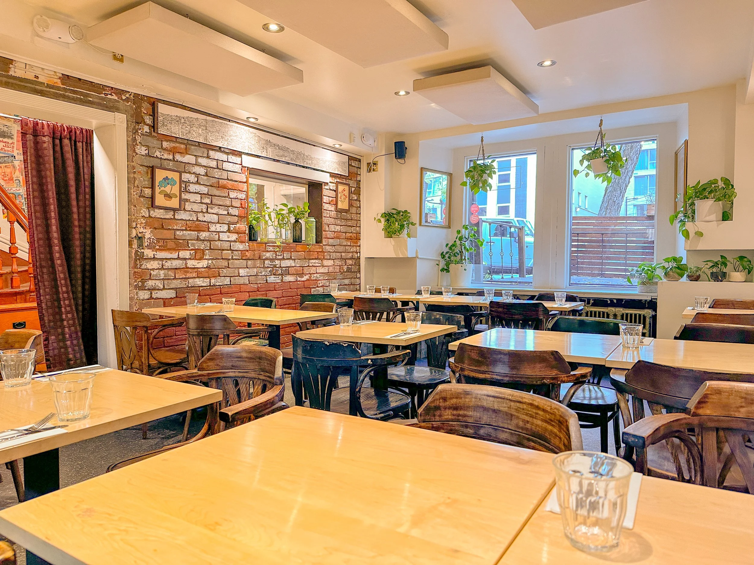 Intérieur d'un restaurant avec plusieurs tables en bois et des chaises en bois foncé. La pièce a un mur en briques apparentes, des plantes vertes sur les fenêtres et des cadres décoratifs. Grandes fenêtres laissant passer beaucoup de lumière naturell