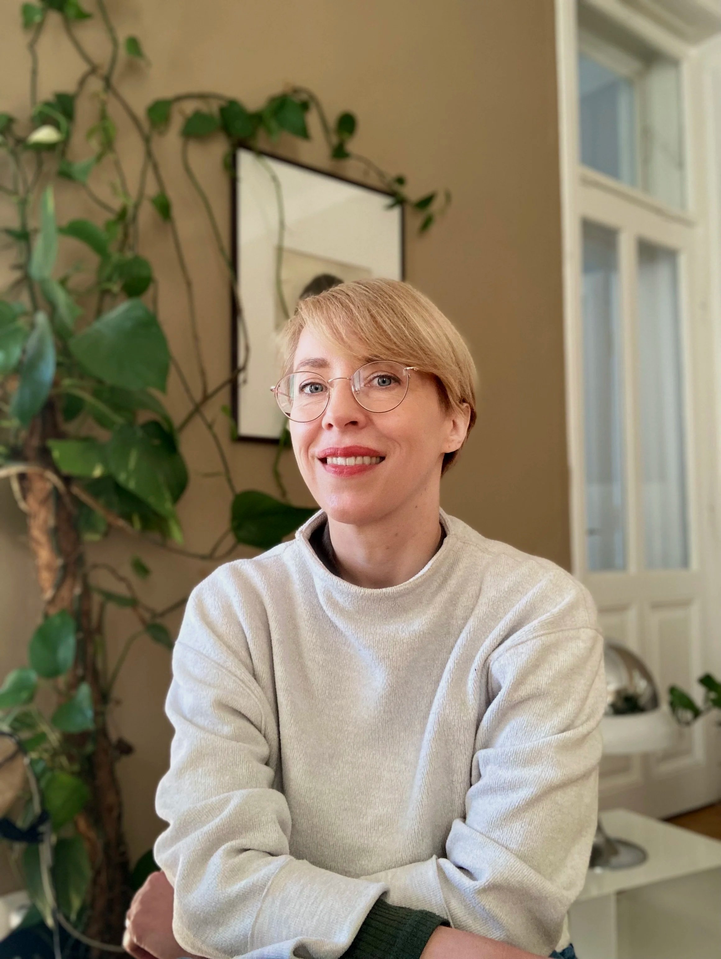 Junge Frau mit kurzen blonden Haaren und Brille, trägt einen hellgrauen Pullover, sitzt vor einer Zimmerpflanze und einem Spiegel in einem hell erleuchteten Raum.