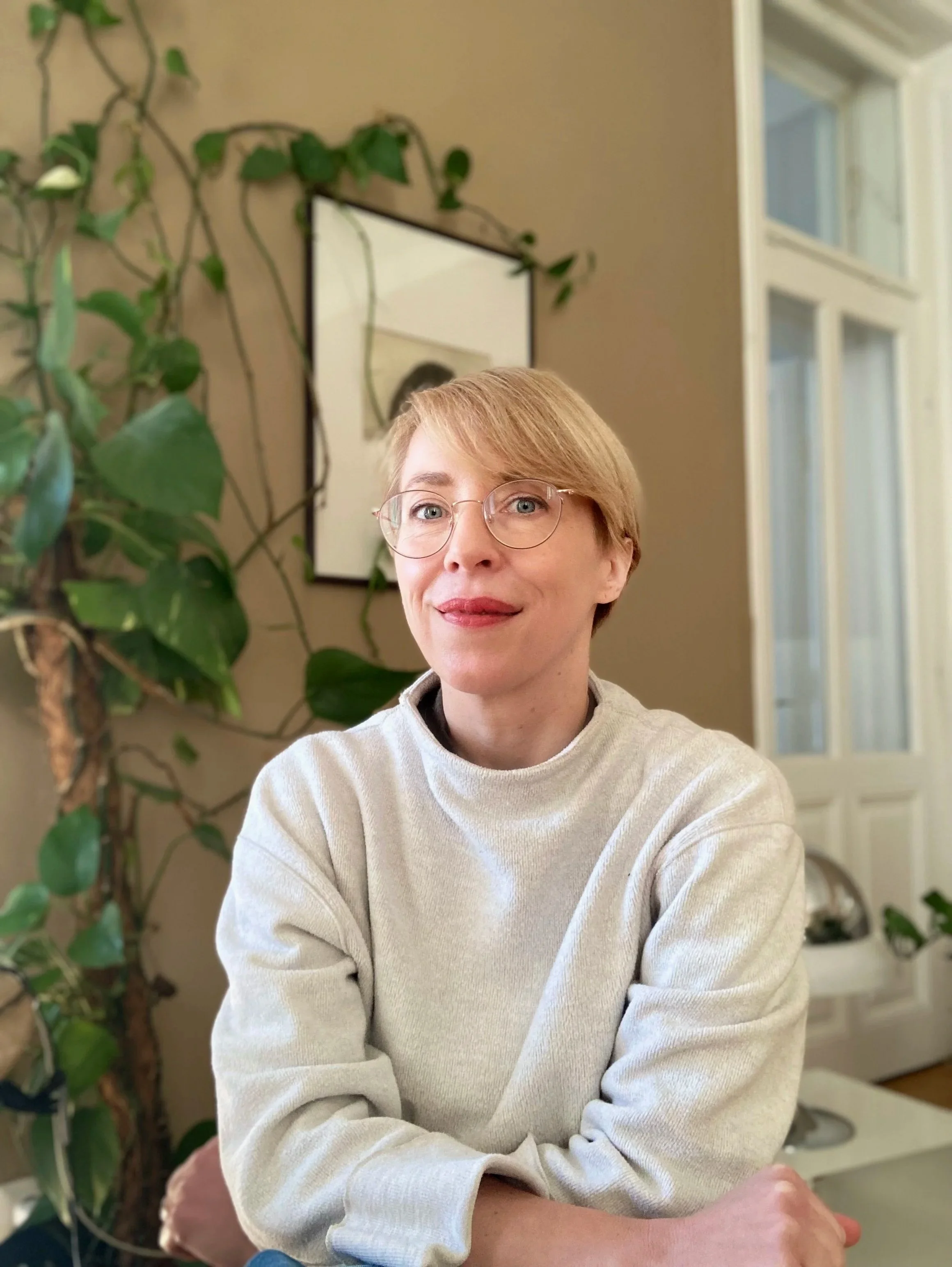 Eine Frau mit kurzen, blonden Haaren und Brille sitzt in einem Raum, hinter ihr eine große Pflanze mit grünen Blättern, ein rechteckiger Spiegel an der Wand und eine weiße Tür mit Fenstern.