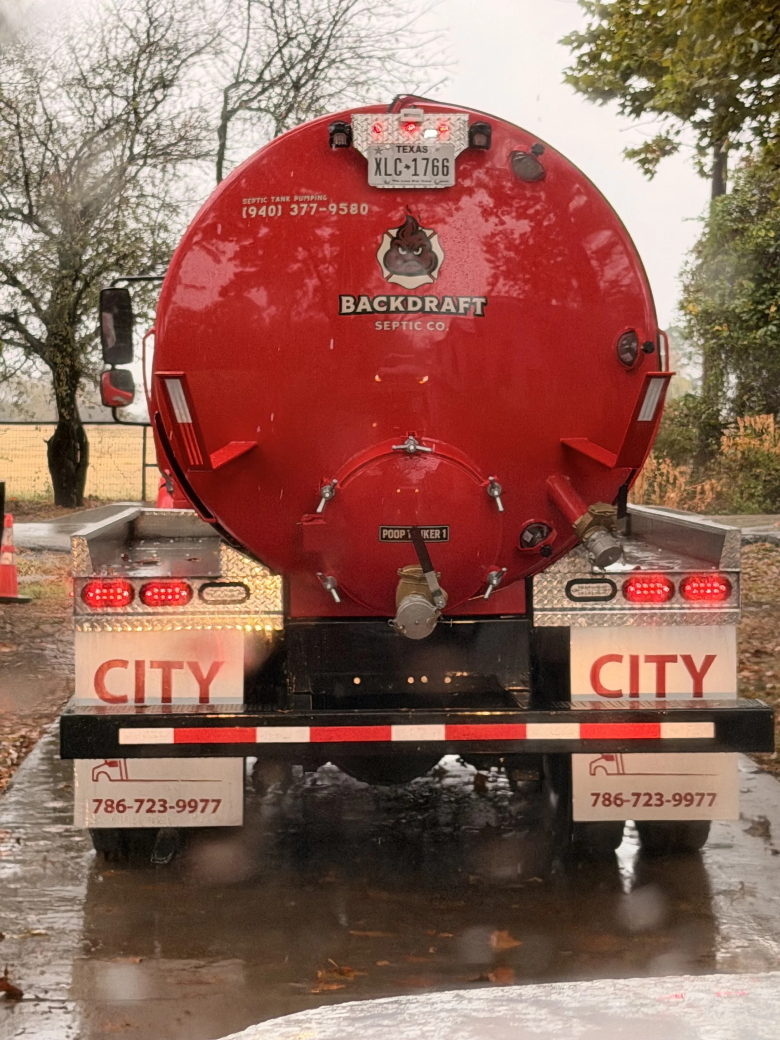 Red septic tank truck with a Backdraft Septic Co. logo featuring an angry poop emoji, parked on a wet road on a rainy day, with trees and a fence in the background.