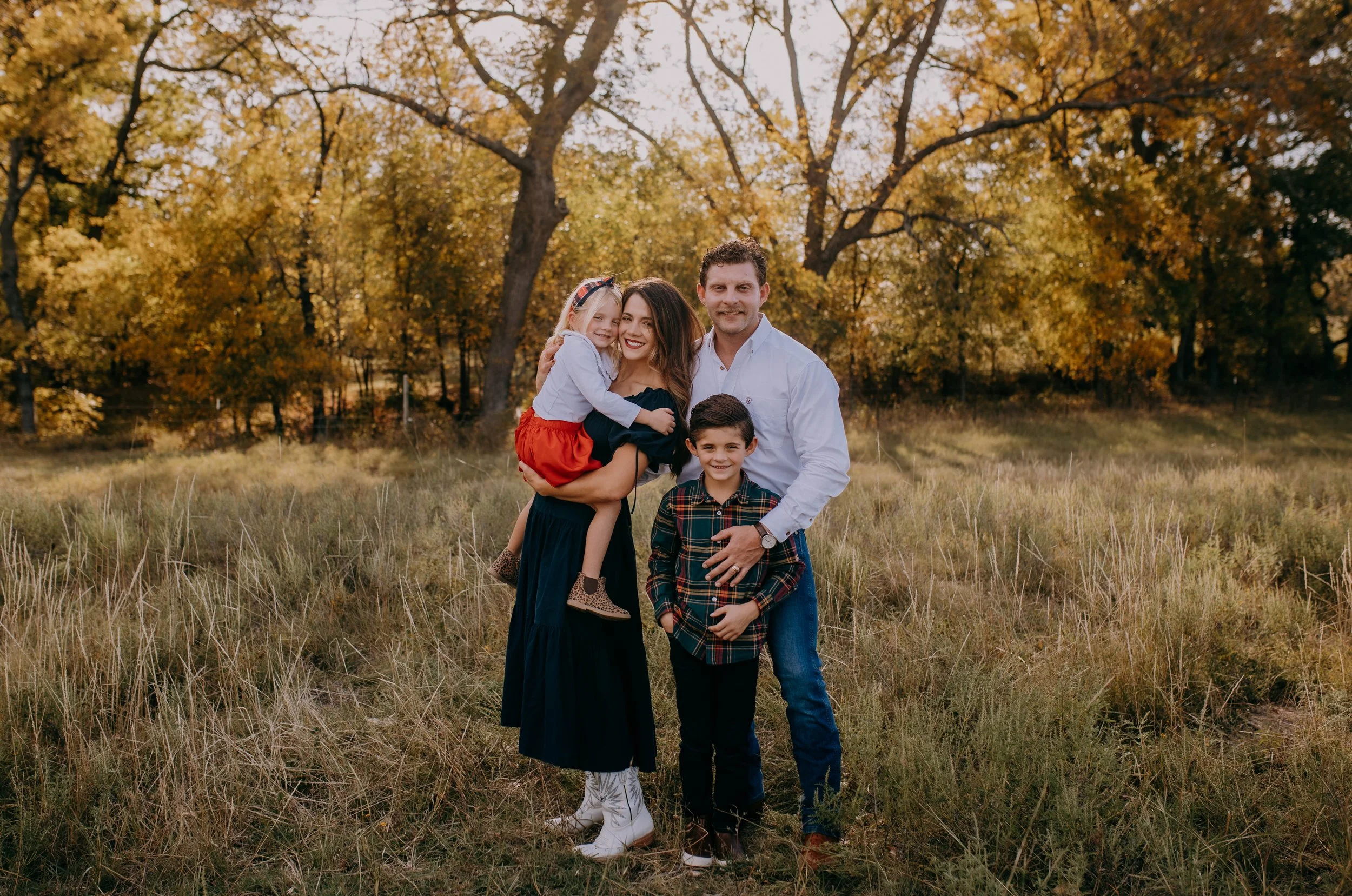A family of four standing outdoors in a field with trees showing fall foliage in the background. The mother holds a young girl, and the father stands beside a young boy. Everyone is smiling and dressed in casual fall clothing.