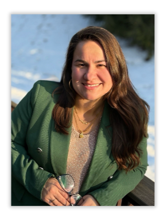 A woman with long brown hair smiling, wearing a green jacket and holding a glass, outdoors in bright sunlight.