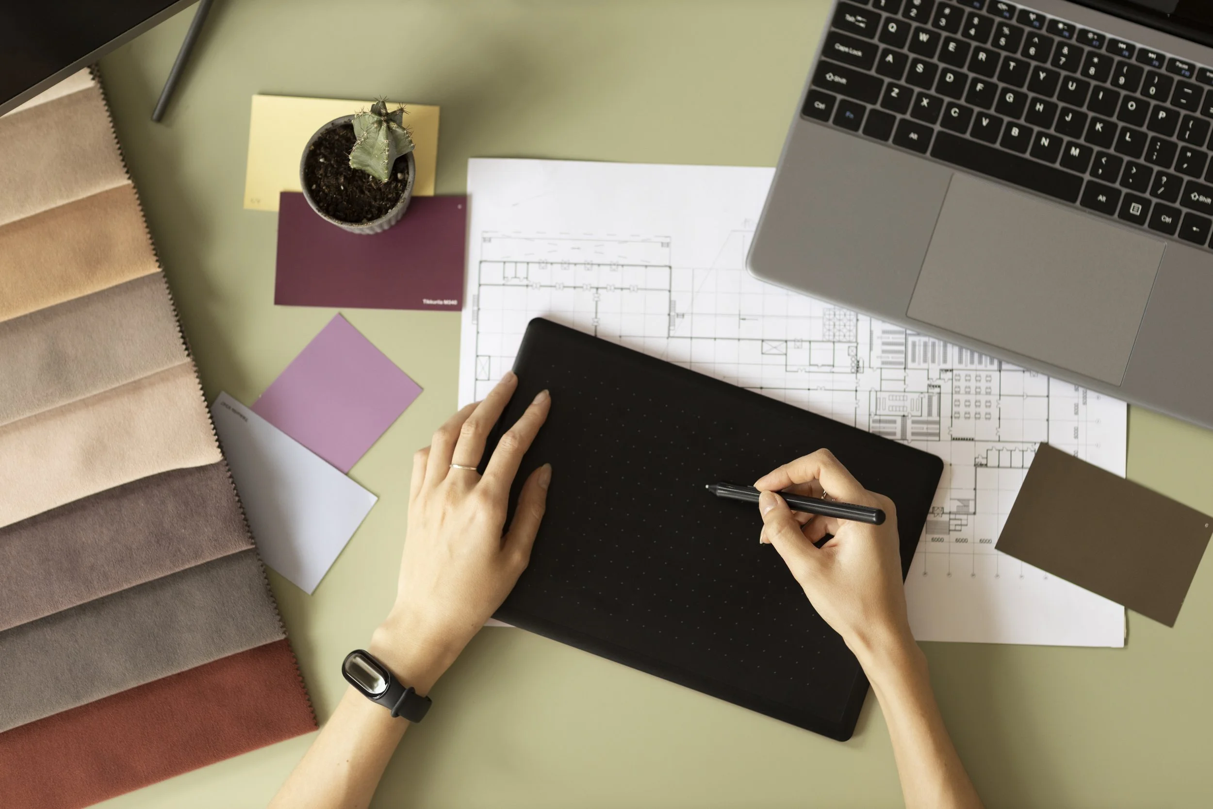 A workspace with a person using a black graphic tablet and stylus, surrounded by color sample swatches, architectural blueprints, a laptop, a small potted cactus, and some color/paint sample sheets on a green desk.