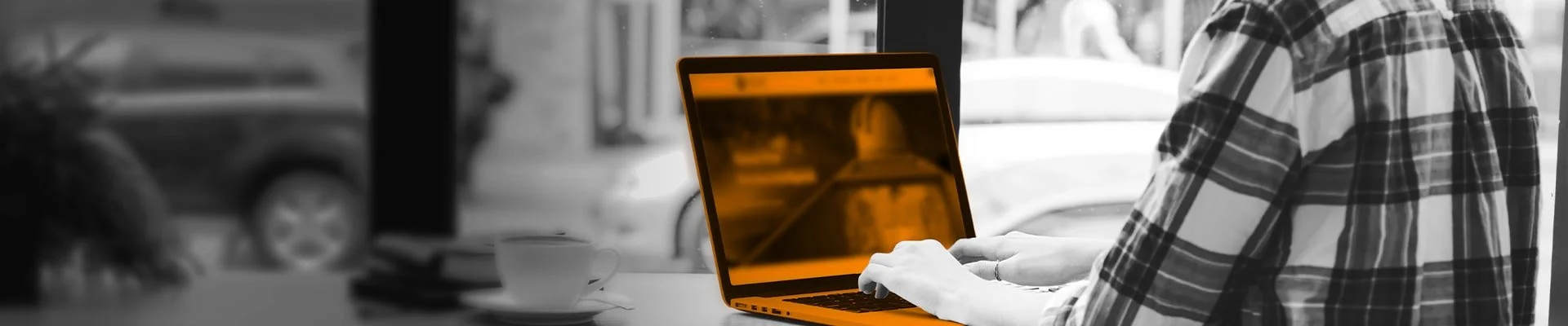 Person using a laptop at a cafe table, with a coffee cup and some books nearby, through a window showing parked cars outside.