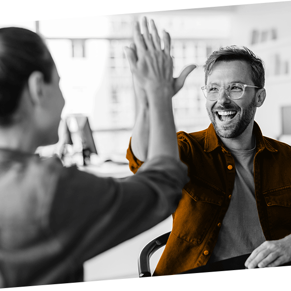 Two people in an office giving each other a high five, with one smiling and wearing glasses and a brown shirt, the other's face not visible.
