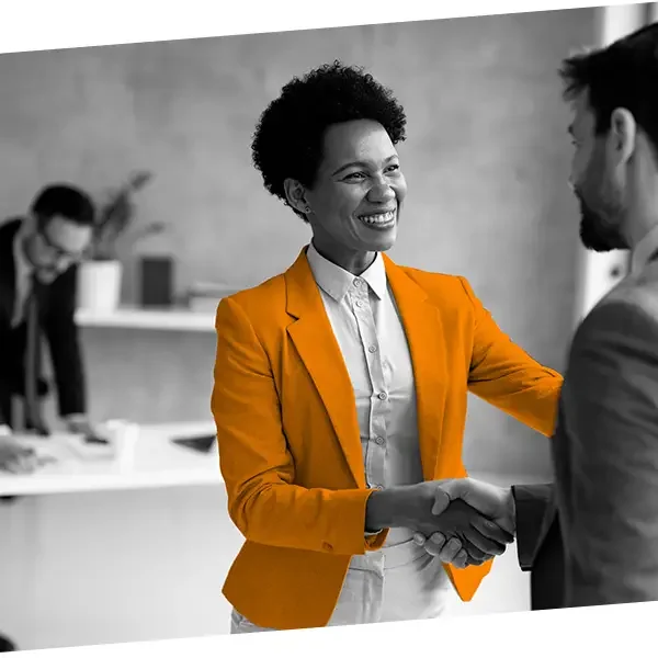 A woman with short curly hair in an orange blazer shaking hands with a man in a suit in an office setting, smiling, with a person in the background working at a desk.