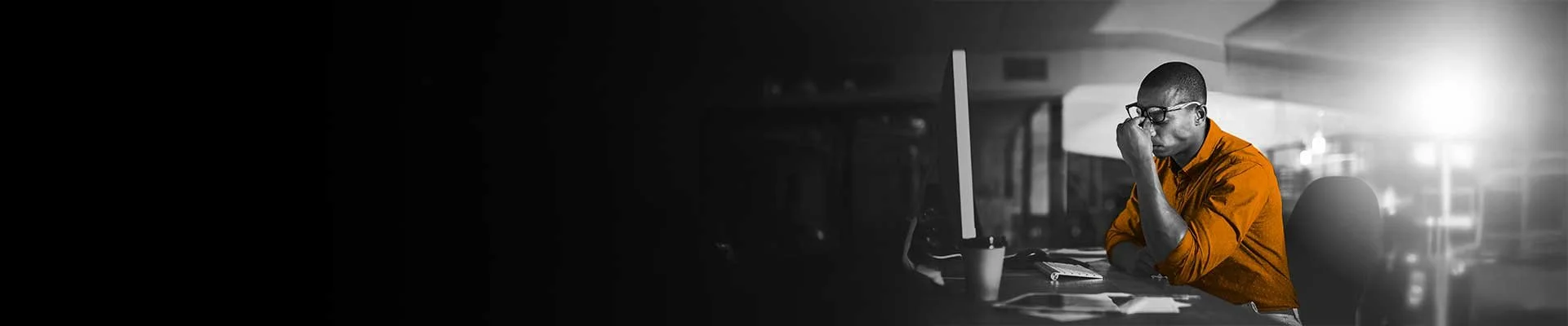 A man wearing glasses and an orange shirt sitting at a desk in front of a computer with a pained or stressed expression, pinching his nose.