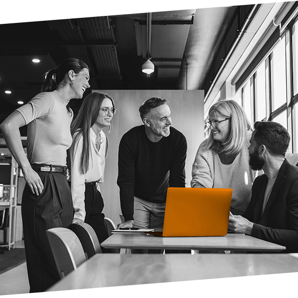 Five diverse professionals in a modern office gathered around an orange laptop, engaging in a collaborative discussion and smiling.