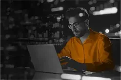 A man wearing glasses and an orange shirt working on a laptop at night in an indoor setting with city lights visible outside