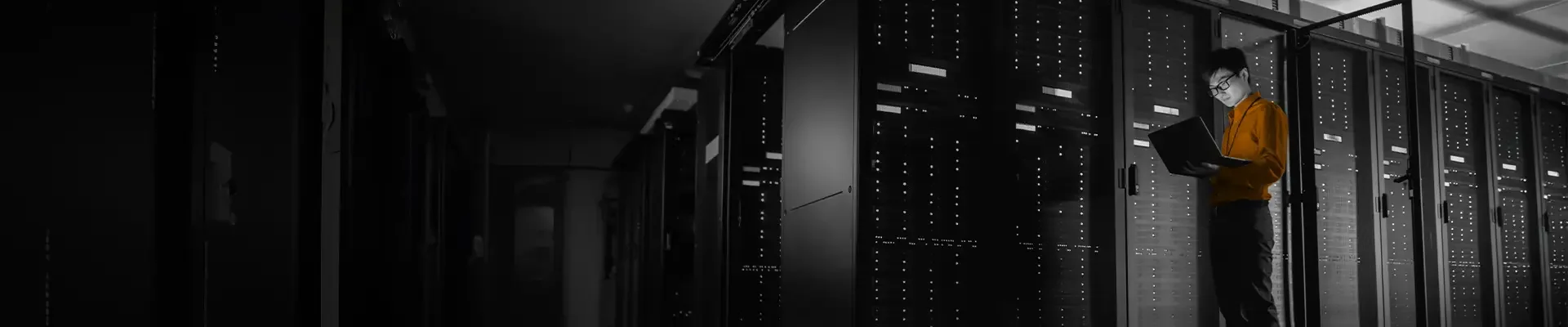 A person wearing glasses in a server room, working on a laptop among rows of server racks.