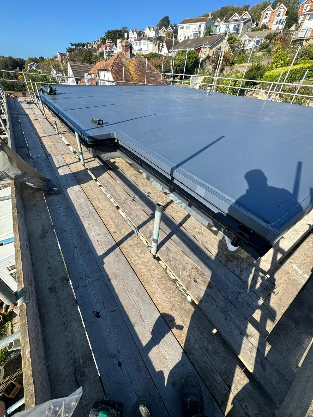 A flat rooftop with solar panels installed, surrounded by safety railings, with a residential neighborhood in the background under a clear blue sky.