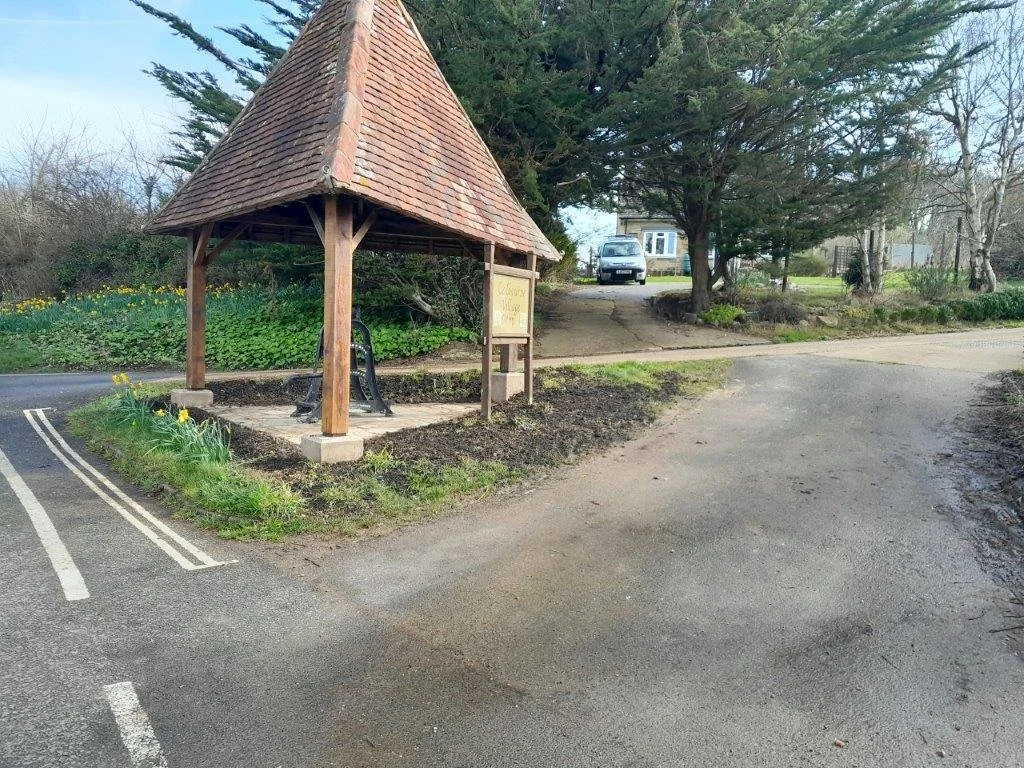 A small wooden shelter with a tiled roof over a bench near a parking area and a dirt walking path, with trees and a house in the background.