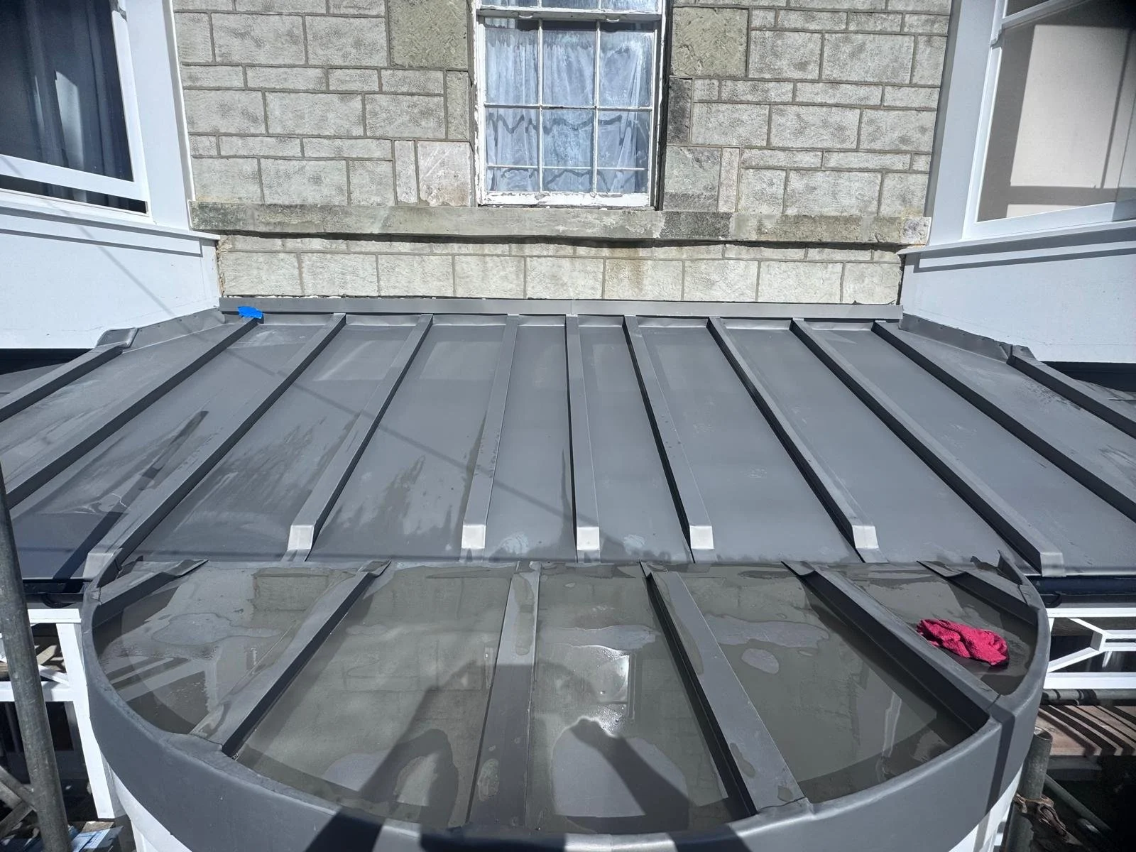 A view from above of a metal awning or roof with a gray color, alongside brick and siding exterior walls of a building, with a window and a pink cloth or towel visible.