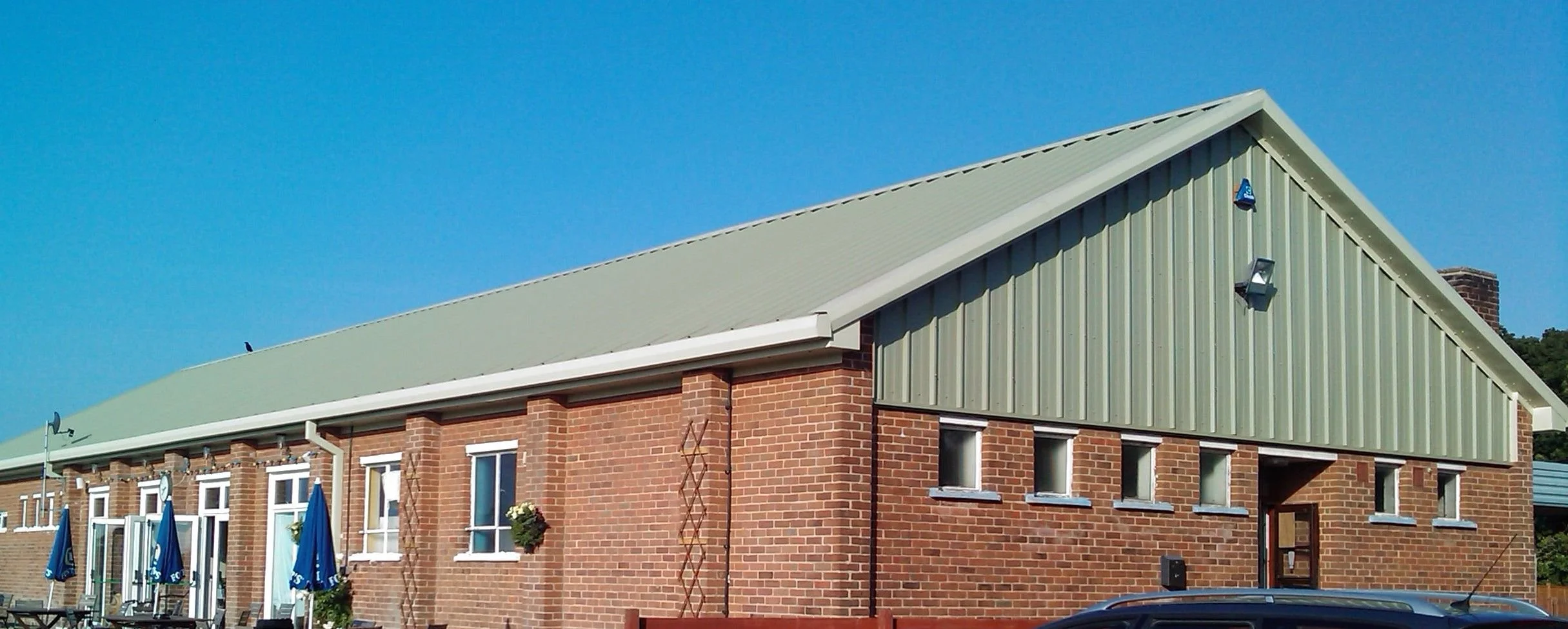 Red brick building with a green sloped metal roof, several small windows, and outdoor seating with blue umbrellas.