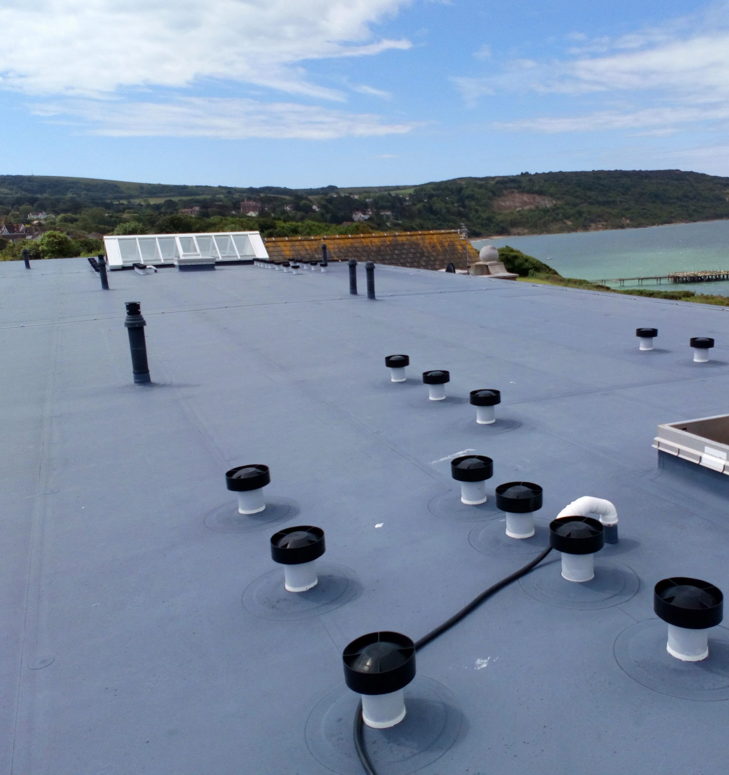 View of a rooftop with multiple vent pipes, a skylight, and a chimney, with a landscape of hills, trees, and houses in the background under a partly cloudy sky.