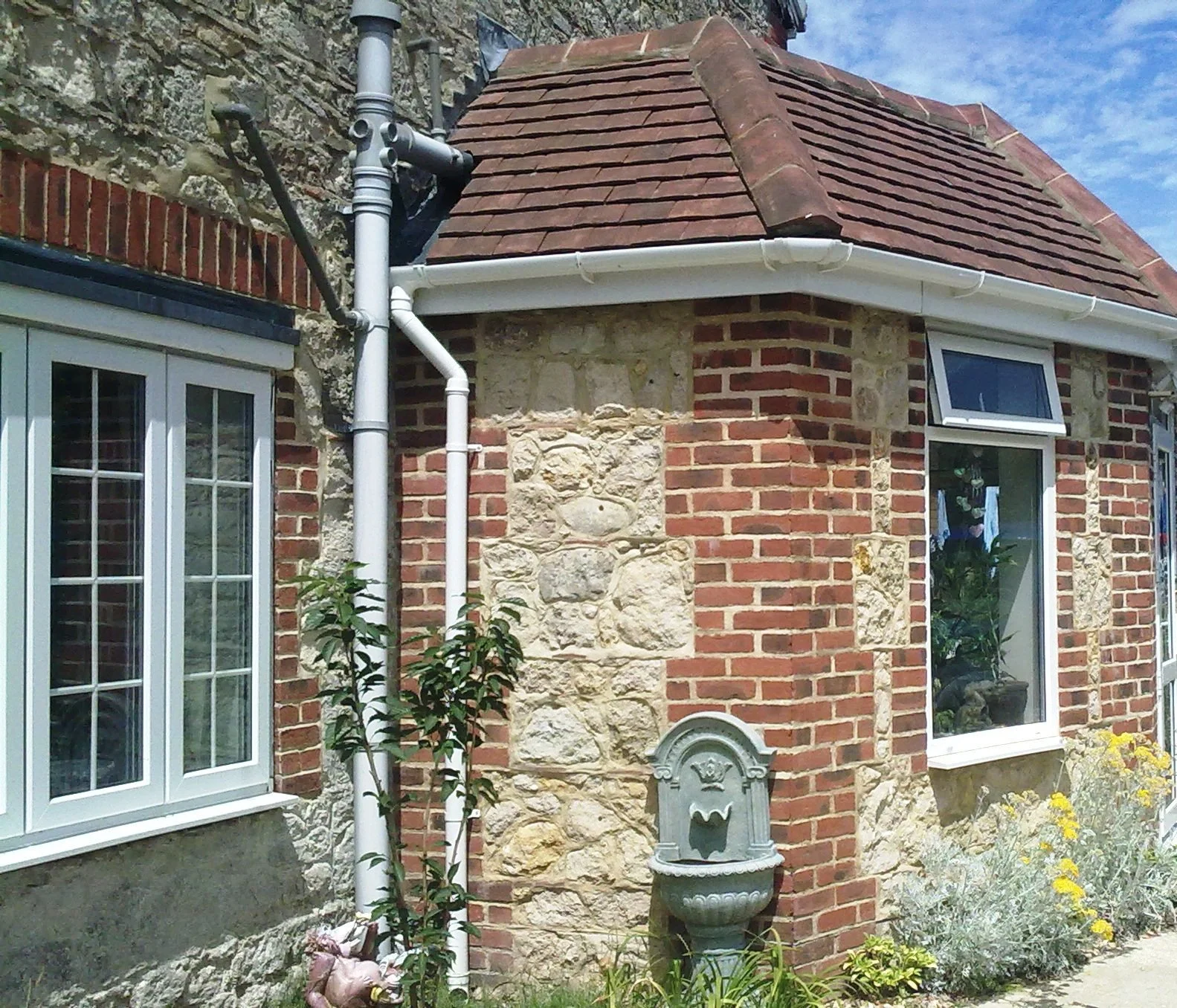 The corner of a brick house with stone accents, a window with white framing, a decorative water fountain, a small plant, and yellow flowers in a garden bed under a blue sky.