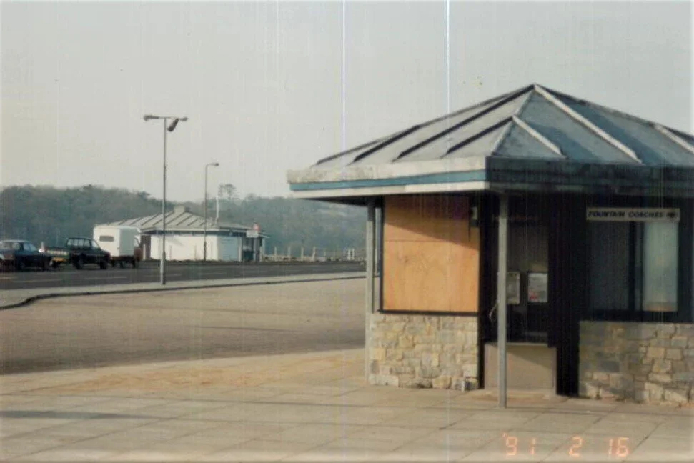 A small, vacant kiosk with a stone base and triangular metal roof, located near a parking lot with several vehicles, and a road with a few streetlights in the background.