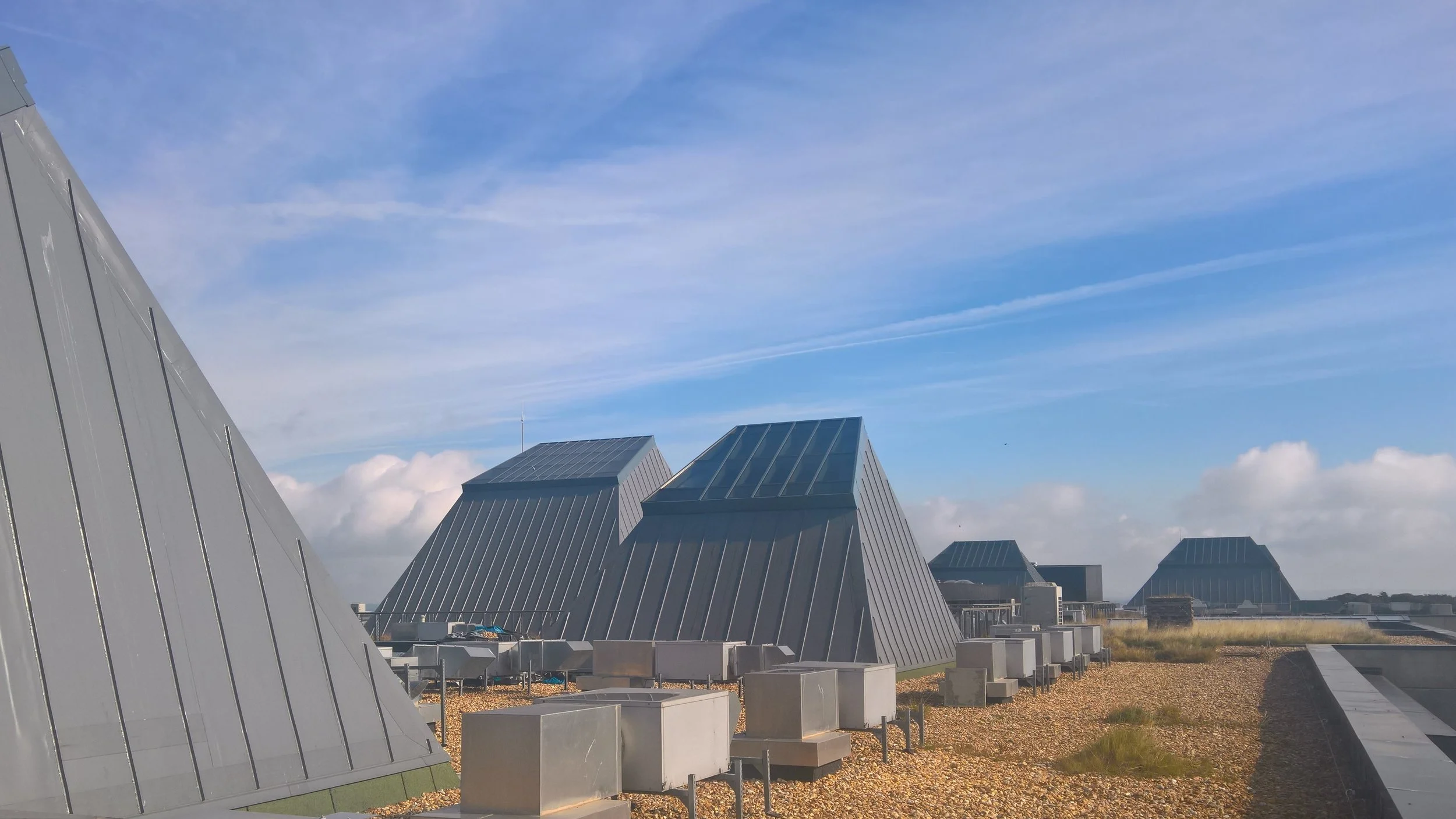 Several modern, metallic, pyramid-shaped buildings with sloped roofs, set against a bright blue sky with streaks of clouds.