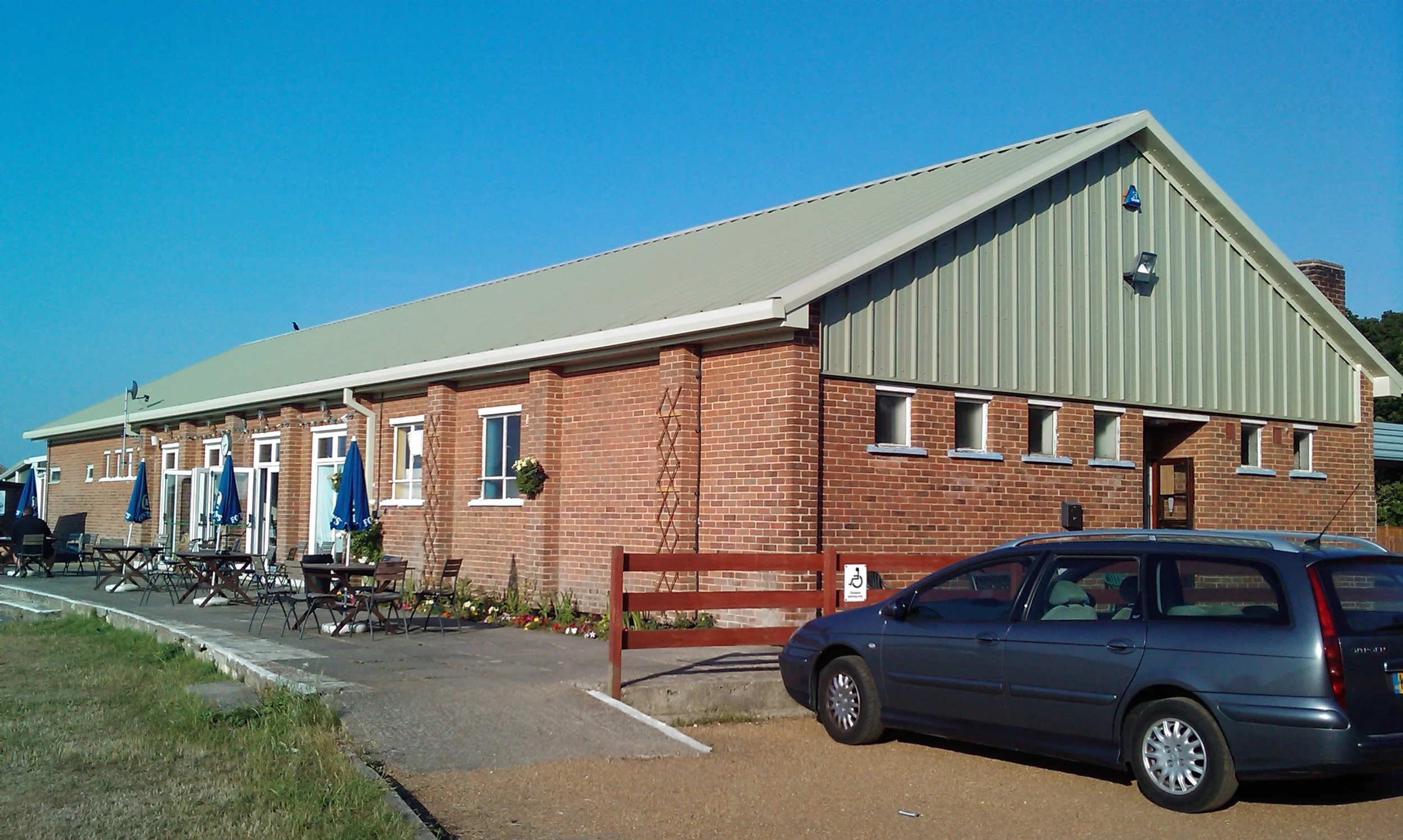 A brick building with a green metal roof, multiple windows, outdoor seating with tables and chairs, and a parking lot with a dark-colored car. Clear blue sky in the background.