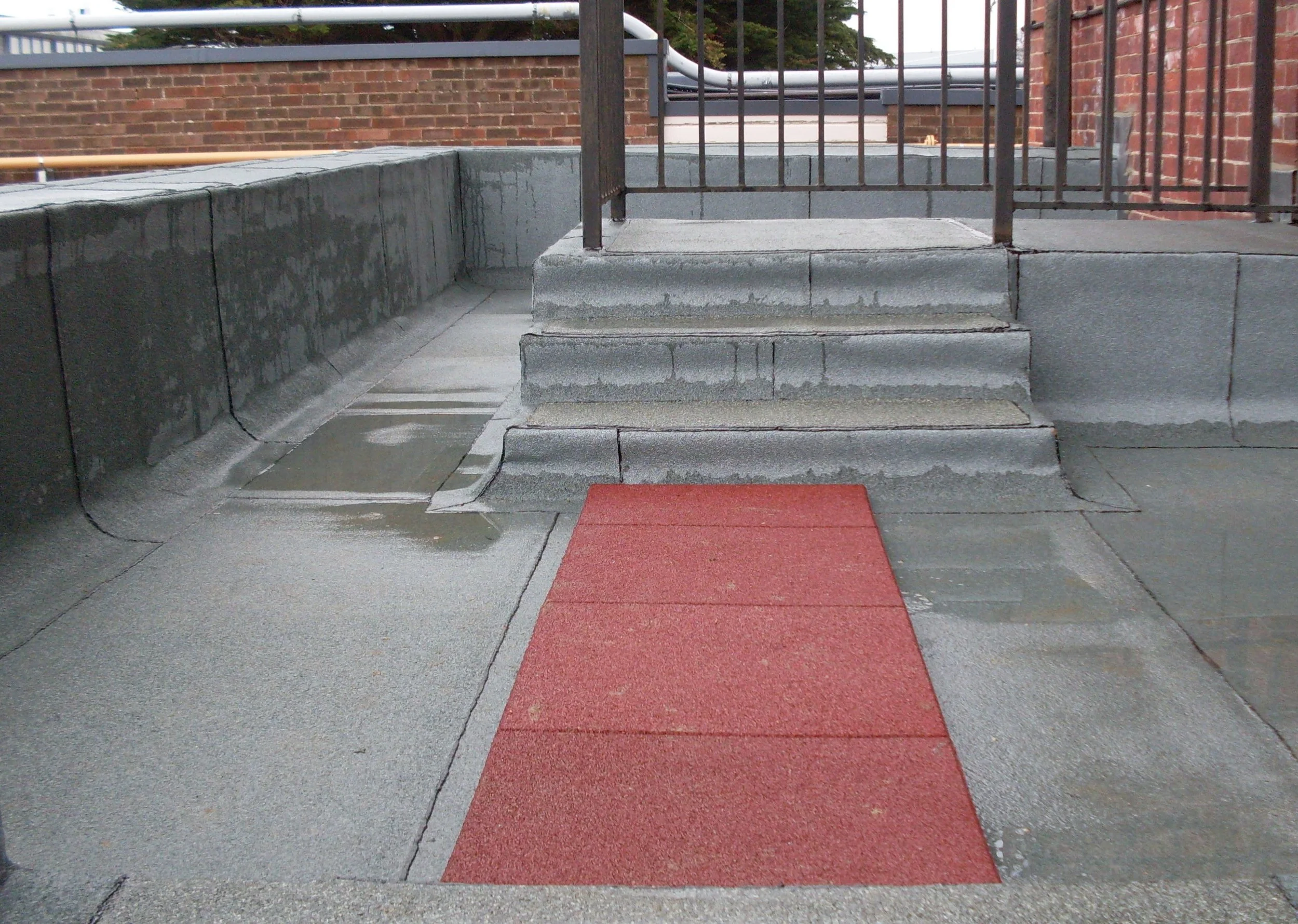 Concrete stairs with three steps, leading to a small landing with a red textured mat. The stairs are adjacent to a concrete wall and a metal railing. In the background, there is a brick wall and some trees.