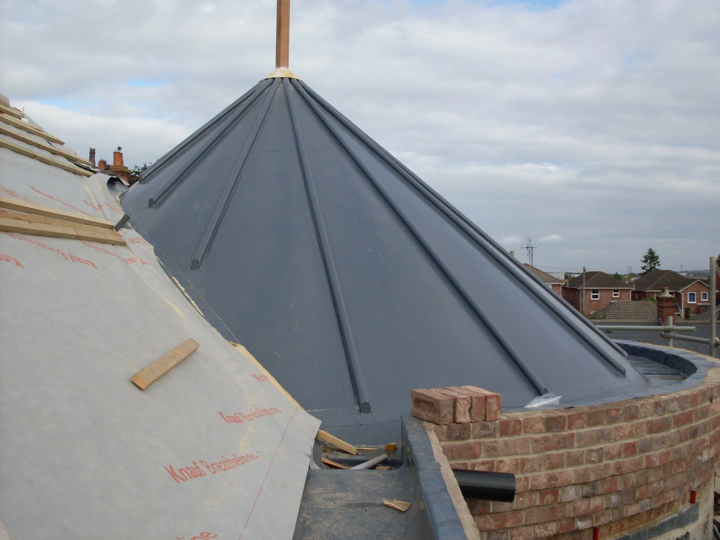 Roof with black metal sheets, brick chimney, and partially completed wooden and insulation layers, with a cloudy sky overhead.
