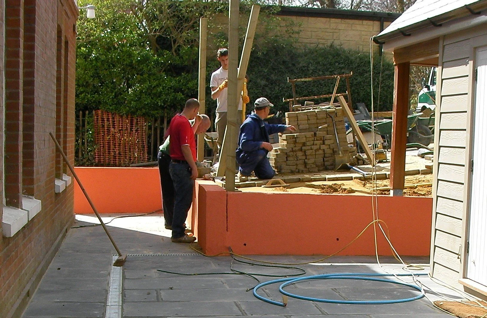 Group of five workers at a construction site, building a brick structure with orange barriers in the background and a white shed on the right side.