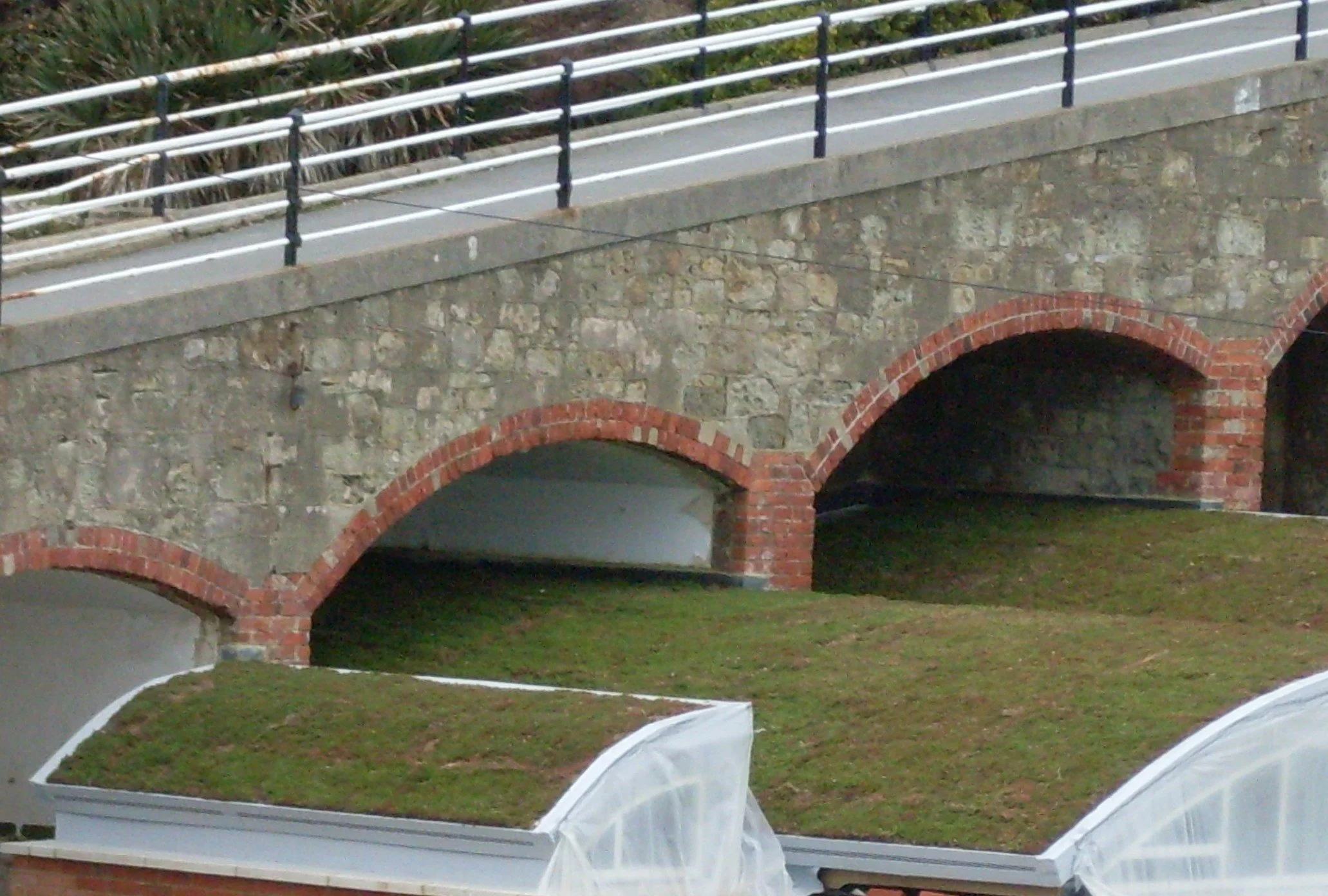 A stone bridge with red brick arches and a metal railing on top, over a grassy area.