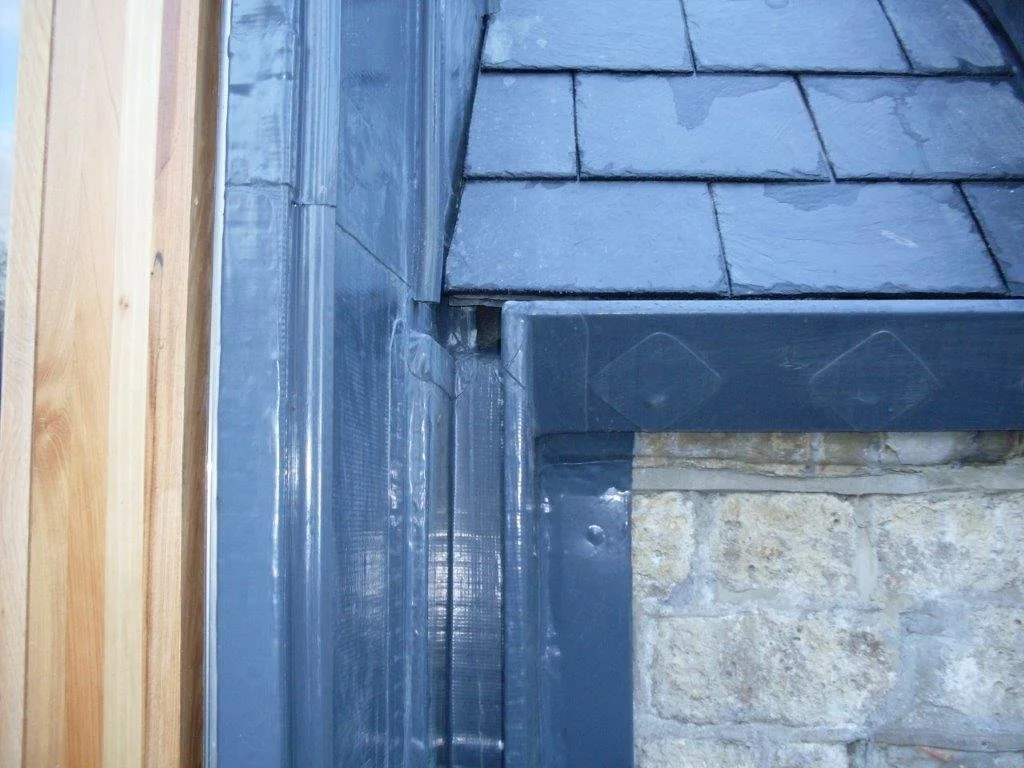 Close-up of a section of a building's exterior, showing a stone wall, a wooden panel, and metal flashing around the roof corner with slate shingles.