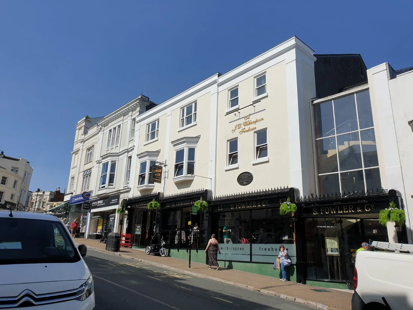 Street scene with white multi-story buildings, storefronts, pedestrians, parked vehicles, and a clear blue sky.