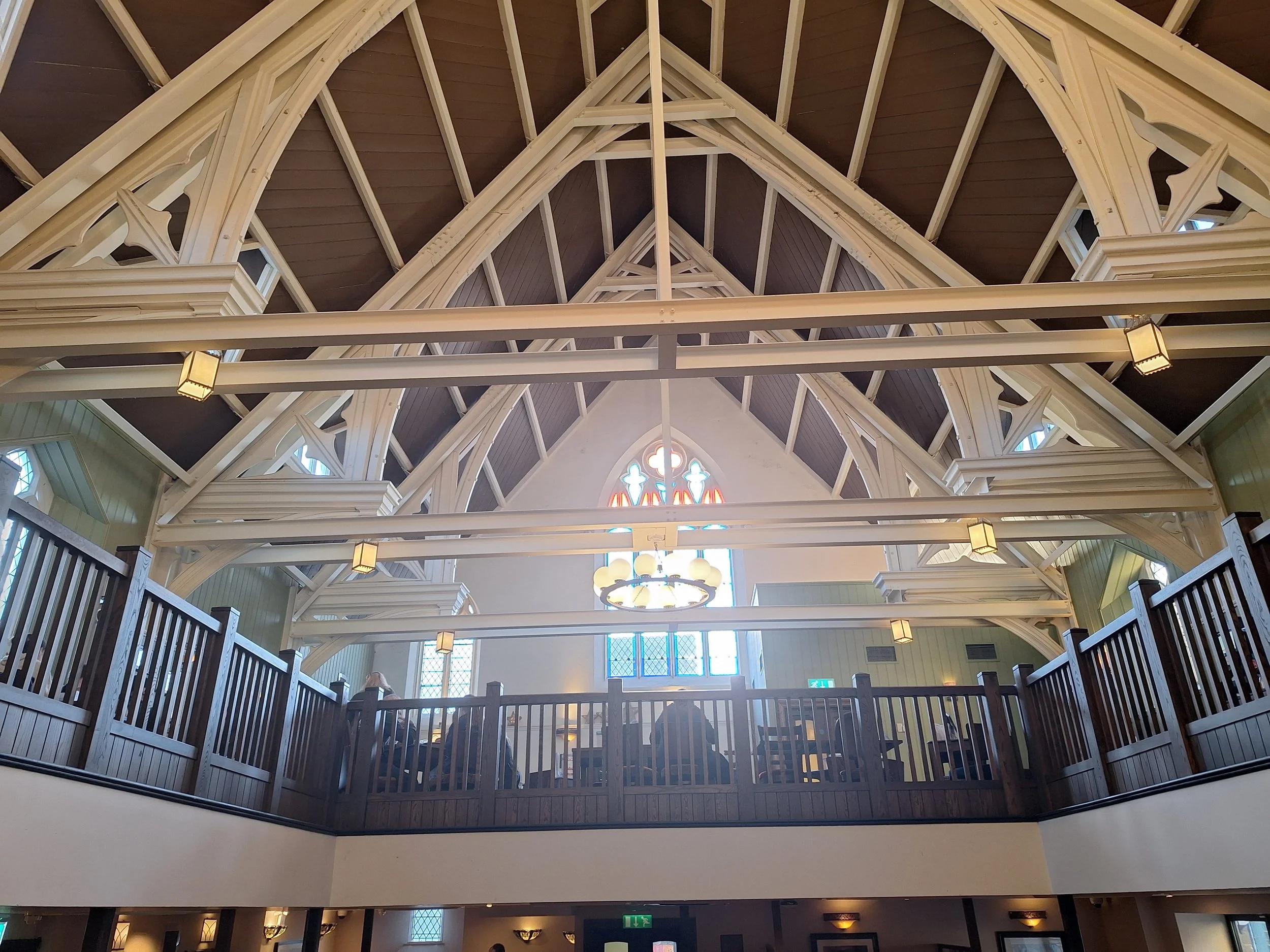 Interior view of a church or chapel with a high wooden vaulted ceiling, stained glass windows, hanging light fixtures, and a balcony with wooden railings.